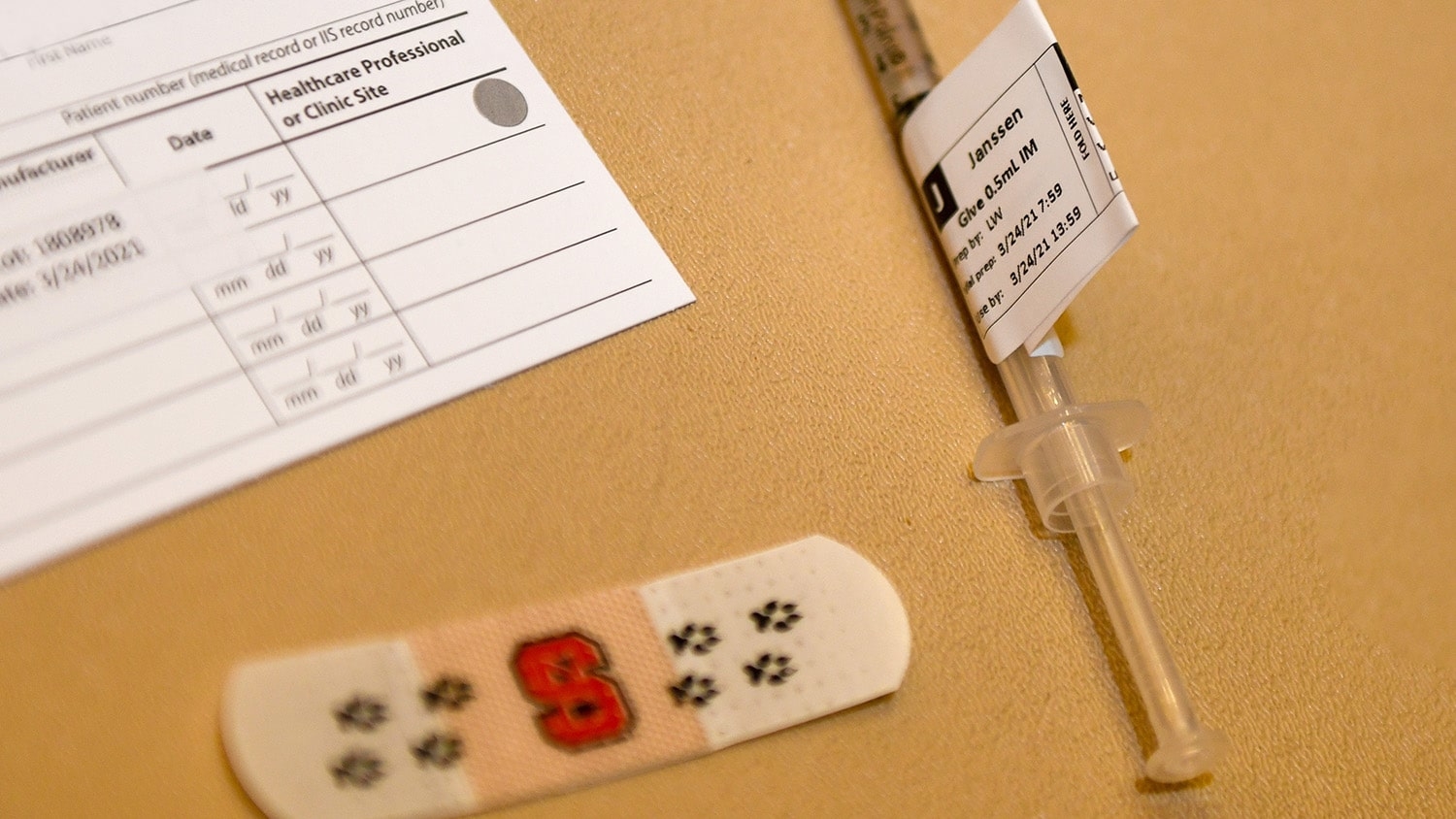 A vaccine card, an NC State-branded bandage and a syringe containing the Janssen vaccine